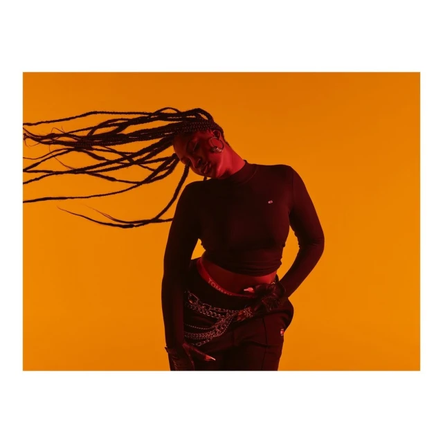 Studio shot of a woman with long braids wearing a black long-sleeved shirt and black pants against a solid orange background.
