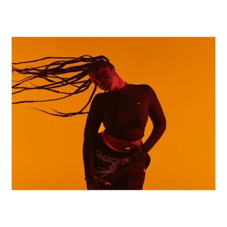 Studio shot of a woman with long braids wearing a black long-sleeved shirt and black pants against a solid orange background.