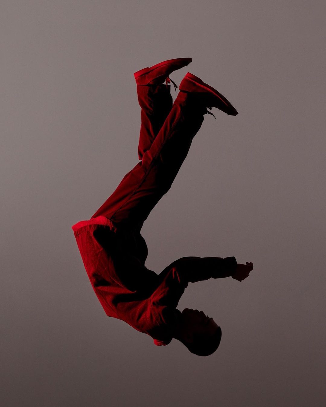 Young African-descent man does acrobatic pose in mid-air wearing red pants and top against a grey background in a studio setting.
