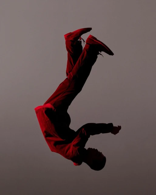 Young African-descent man does acrobatic pose in mid-air wearing red pants and top against a grey background in a studio setting.