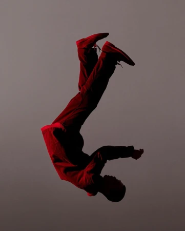 Young African-descent man does acrobatic pose in mid-air wearing red pants and top against a grey background in a studio setting.