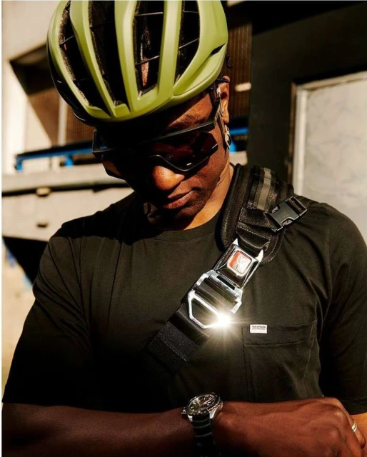 Adult male cyclist wearing a helmet and carrying a sling bag, looking down at his wrist watch in an urban environment.