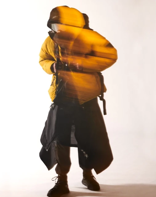 Person wearing a yellow puffer jacket and black draped pants in a studio shot with a blurred motion effect, creating a dynamic and modern fashion image.