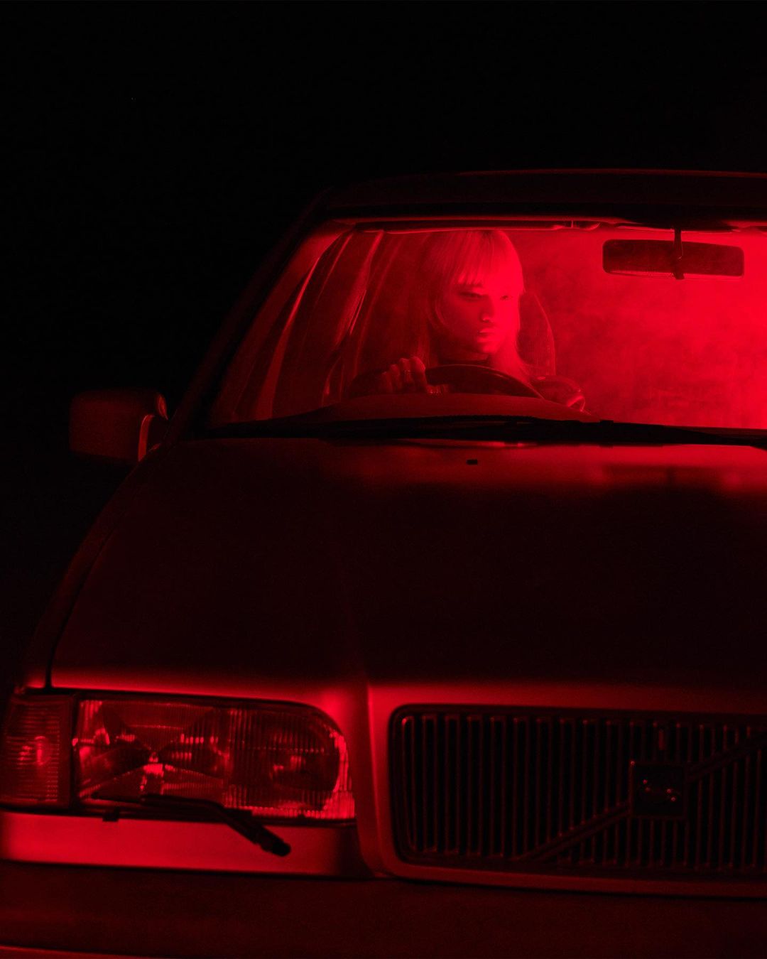 Red-lit car interior featuring a woman in the driver's seat, creating a striking and mysterious visual composition in dark environment.