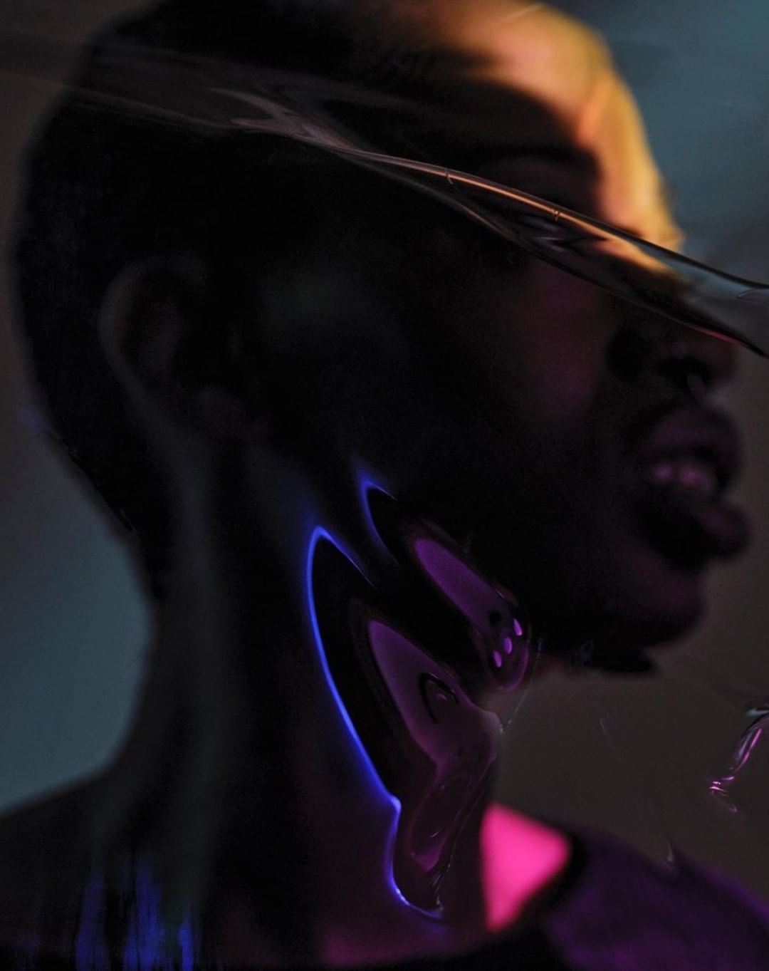 Dramatic portrait of a Black adult male's face partially obscured by a distorted plastic sheet, featuring vibrant neon pink and blue lighting.