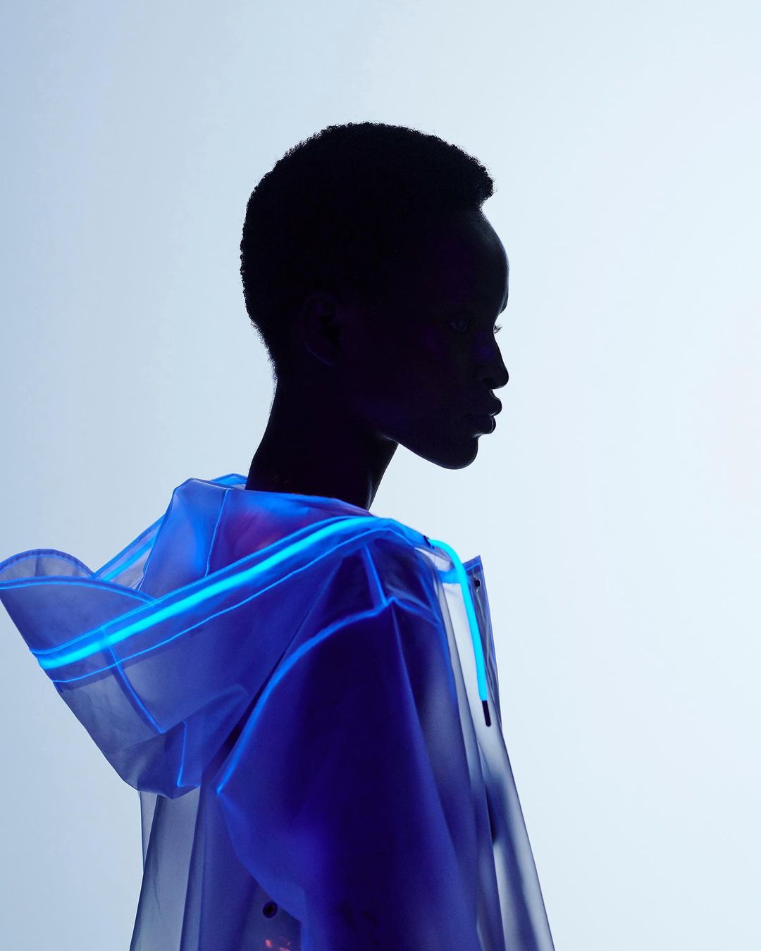 Profile shot of a silhouetted figure in a translucent raincoat with blue neon piping against a gradiated light blue backdrop.