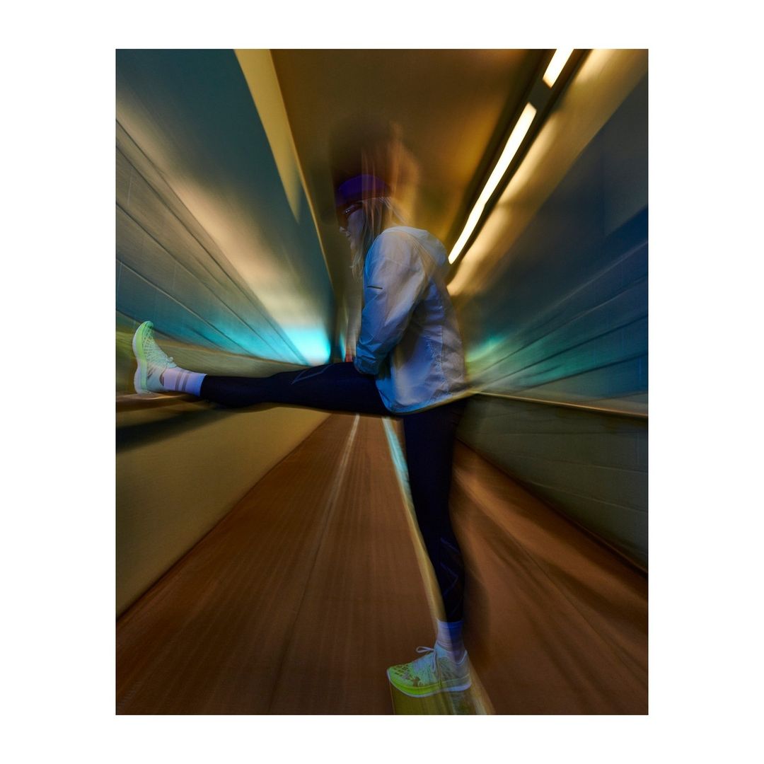 A woman in athletic clothing stretches her leg in a tunnel with directional lighting, creating a dynamic, motion-blurred fitness image.