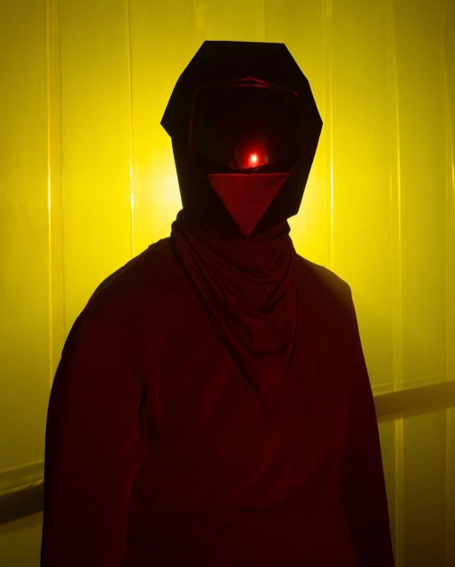Person in a dark hood with a red light in a geometric helmet stands against a yellow panel wall in an indoor setting.
