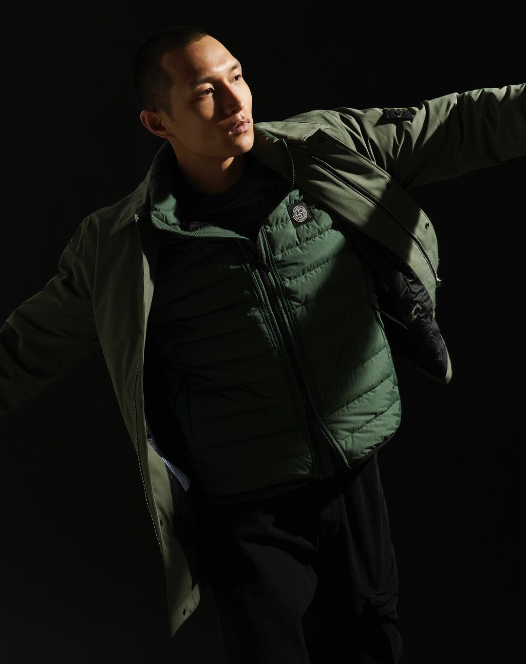 Adult male fashion model poses in olive green jacket and vest against a dark background in a studio setting with dramatic lighting.
