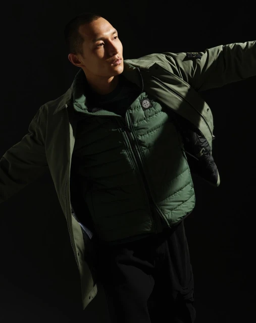 Adult male fashion model poses in olive green jacket and vest against a dark background in a studio setting with dramatic lighting.