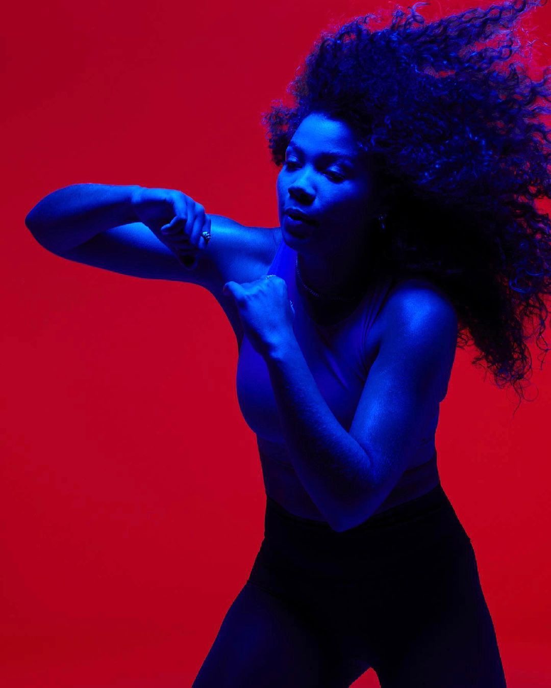 Woman in fitness attire strikes a boxing pose against a vivid red backdrop, illuminated by blue lighting, showcasing athletic form and dynamic movement.