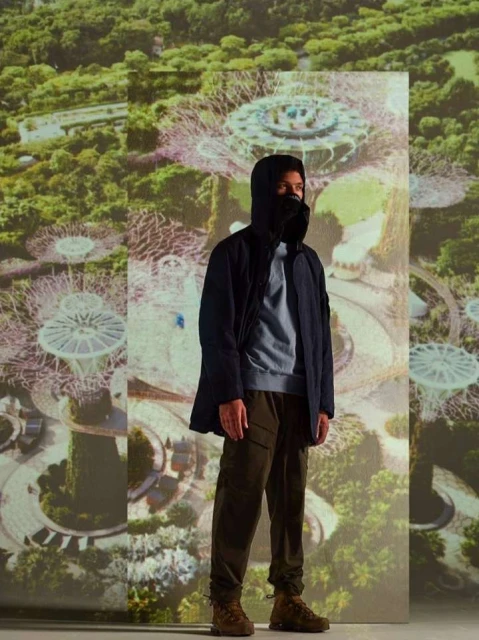 A man in outdoor wear poses in front of a projected image of an aerial cityscape, modelling outdoor jacket, sweatshirt, trousers and boots.