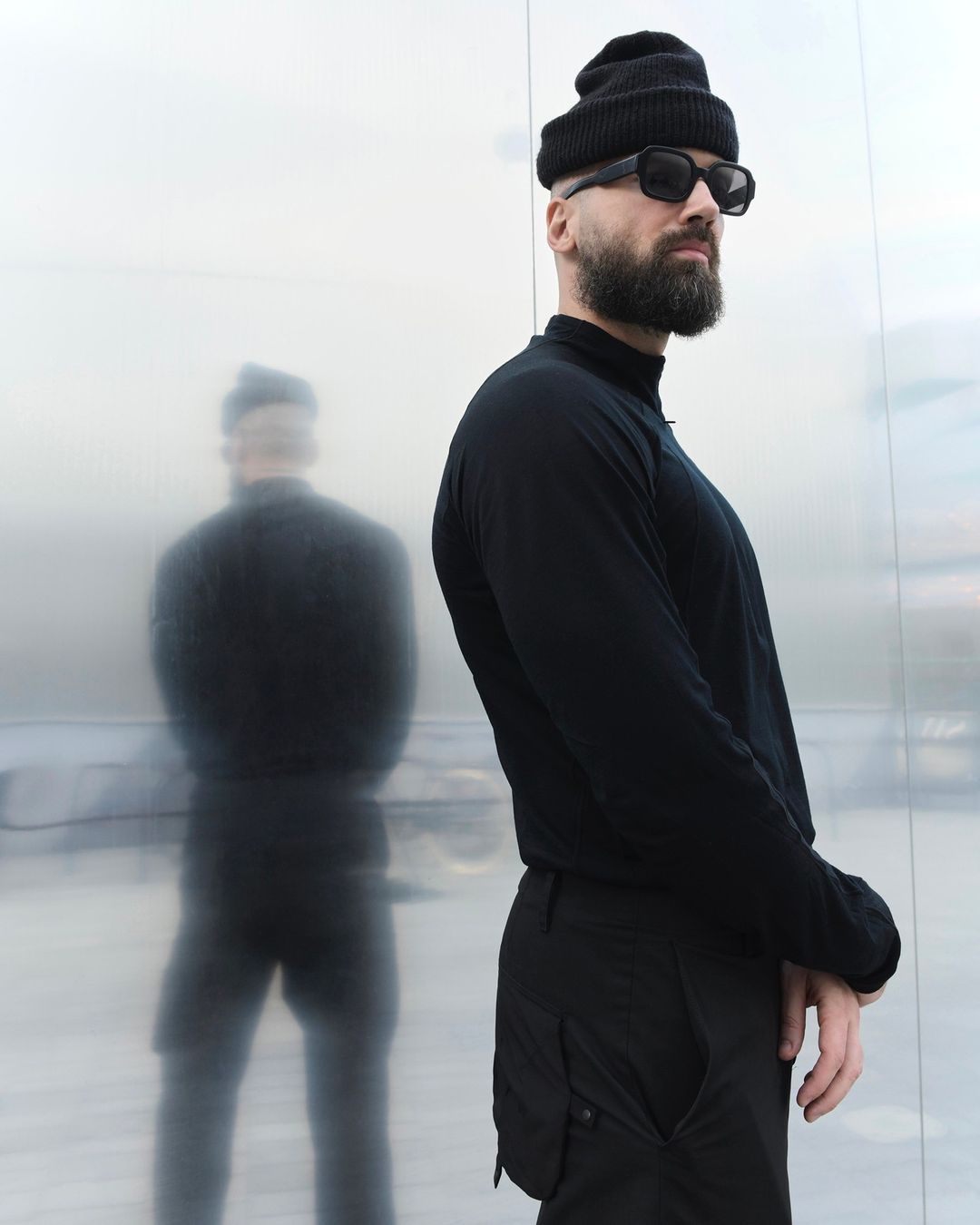 Adult male wearing a black beanie, long-sleeve shirt, and sunglasses stands in front of a reflective wall, with a blurred shadow figure.