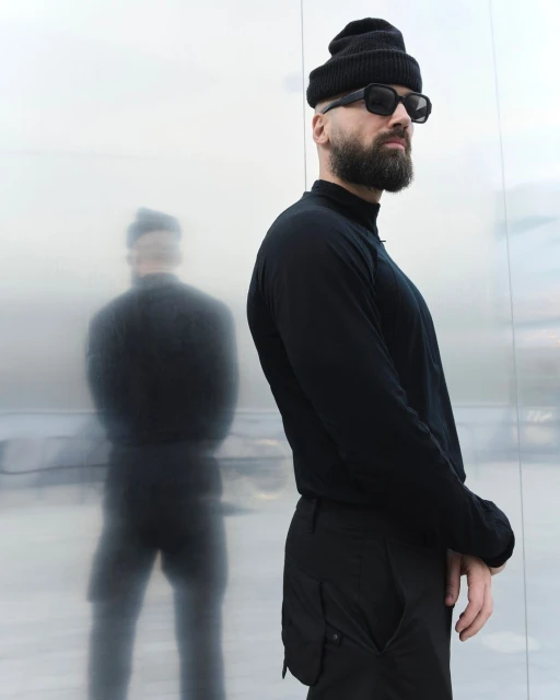 Adult male wearing a black beanie, long-sleeve shirt, and sunglasses stands in front of a reflective wall, with a blurred shadow figure.