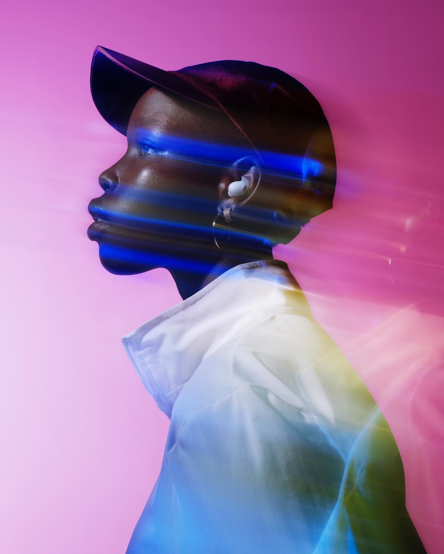 Profile view of an adult, with a blurred light effect, wearing a white jacket and black cap against a pink background in a studio setting.