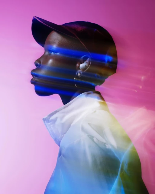 Profile view of an adult, with a blurred light effect, wearing a white jacket and black cap against a pink background in a studio setting.