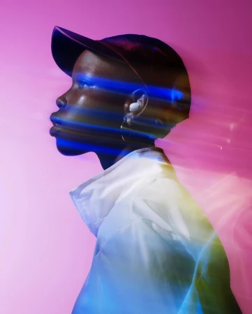 Profile view of an adult, with a blurred light effect, wearing a white jacket and black cap against a pink background in a studio setting.