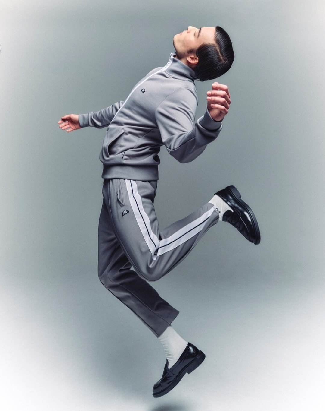 Man in a grey tracksuit and black leather loafers mid-jump against a gradient grey background in a studio fashion shot.