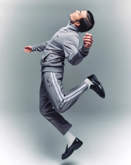 Man in a grey tracksuit and black leather loafers mid-jump against a gradient grey background in a studio fashion shot.