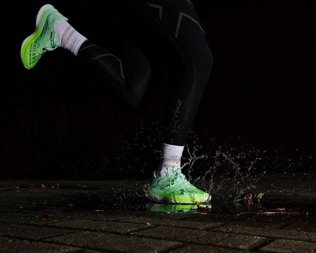 Close-up of a runner's foot landing in a puddle, splashing water around a neon green running shoe and black athletic tights.