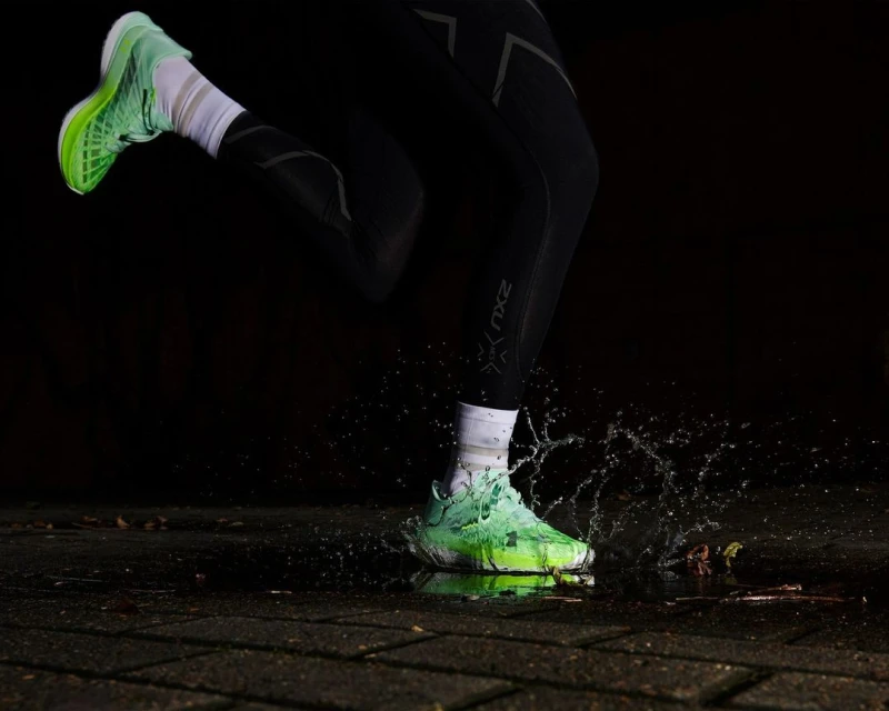Close-up of a runner's foot landing in a puddle, splashing water around a neon green running shoe and black athletic tights.