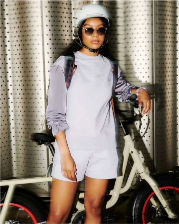 Woman in helmet poses next to a bicycle in studio. She wears a matching lilac sweat suit and sunglasses with a decorative backdrop.