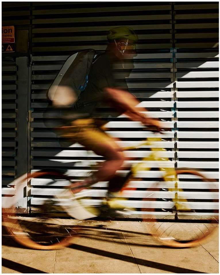 Motion blur of a cyclist wearing a helmet and backpack riding a bike past a wall with horizontal lines, creating dynamic shadows.