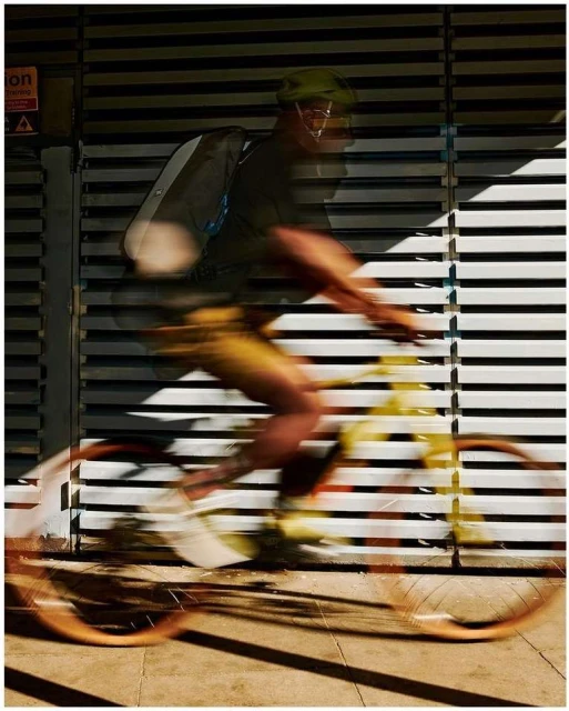 Motion blur of a cyclist wearing a helmet and backpack riding a bike past a wall with horizontal lines, creating dynamic shadows.