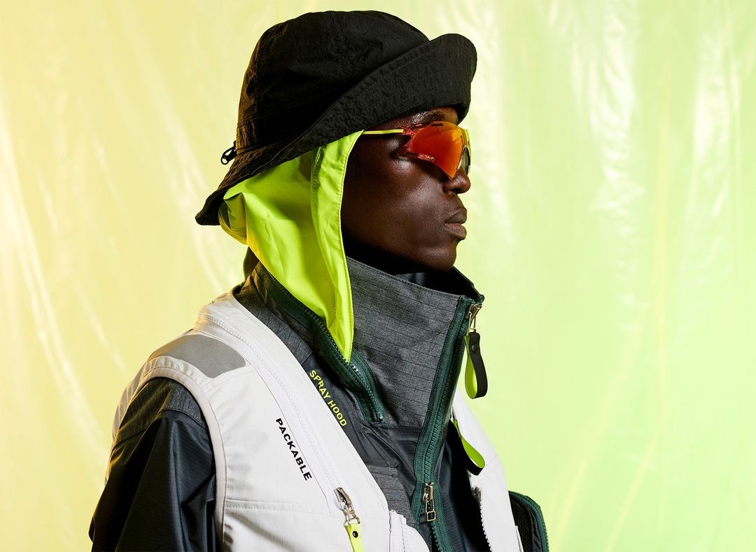 Profile of a Black adult male wearing sunglasses, a vest, and a bucket hat with a neon green hood over a vibrant lime green background.