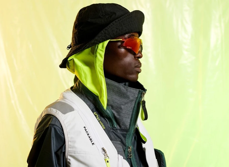 Profile of a Black adult male wearing sunglasses, a vest, and a bucket hat with a neon green hood over a vibrant lime green background.