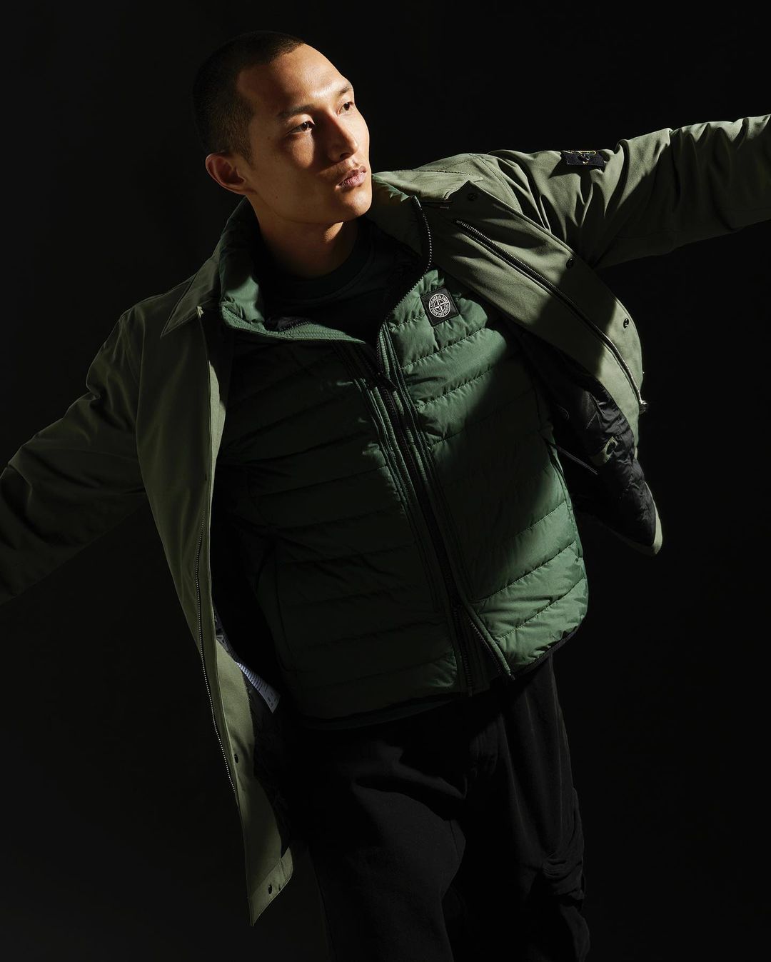 Adult male in a green quilted jacket and light green overshirt poses in front of a dark backdrop, arms slightly extended.