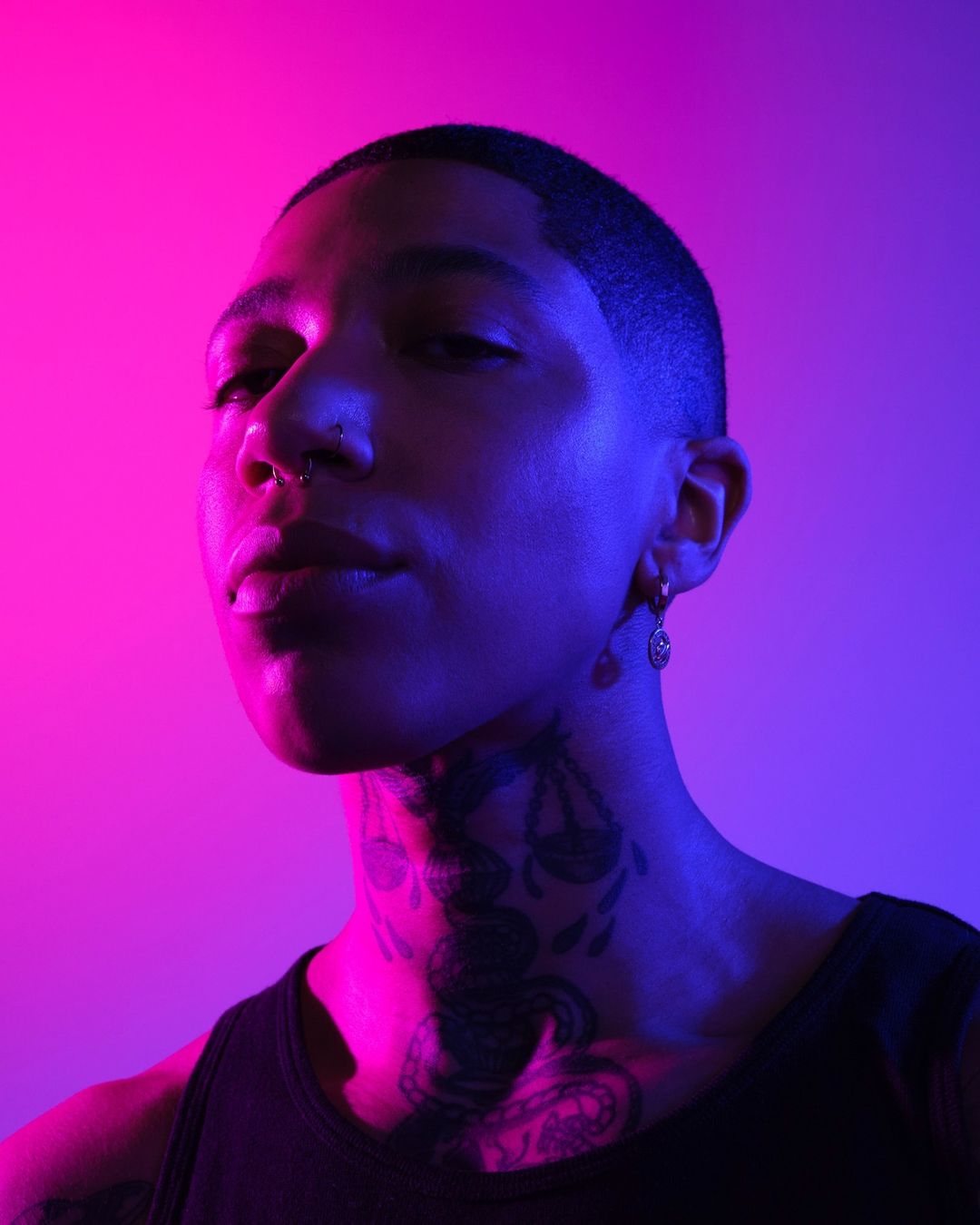 Close-up studio portrait of a young adult with a neck tattoo, adorned with piercings and earrings, set against a vibrant pink and purple backdrop.