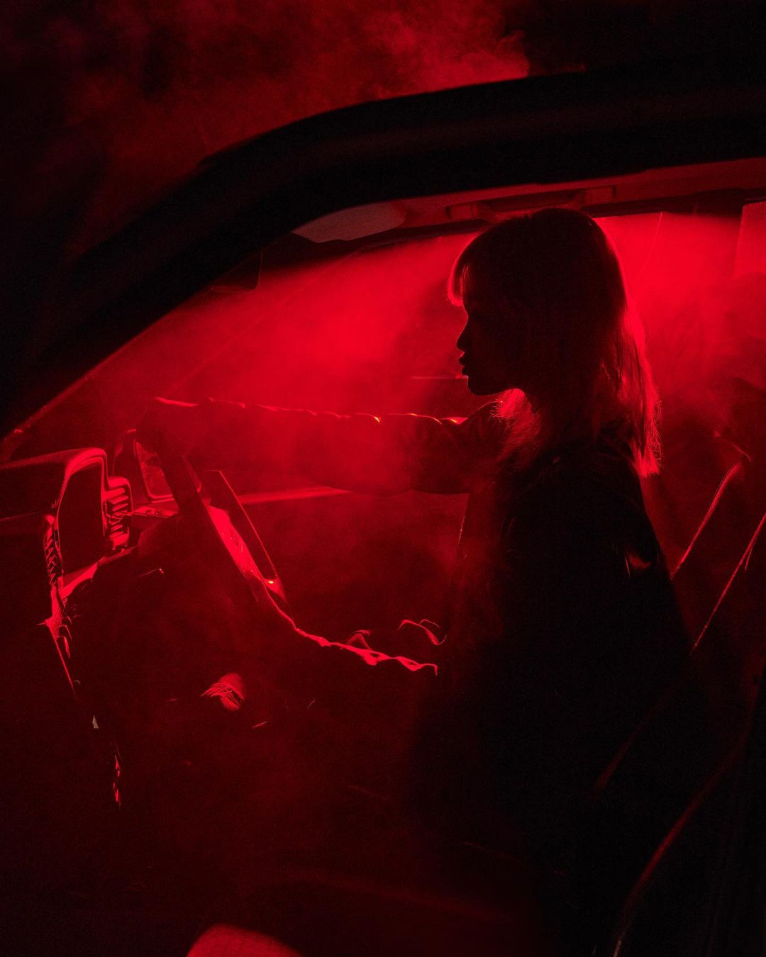 Silhouette of a woman behind the steering wheel of a car, bathed in intense red light. The scene has an edgy, dramatic atmosphere.