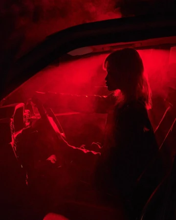 Silhouette of a woman behind the steering wheel of a car, bathed in intense red light. The scene has an edgy, dramatic atmosphere.