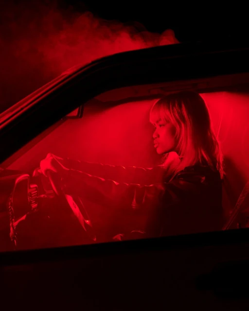 Woman driving car at night under red lighting. The vehicle's interior is dark, with a moody, cinematic aesthetic.