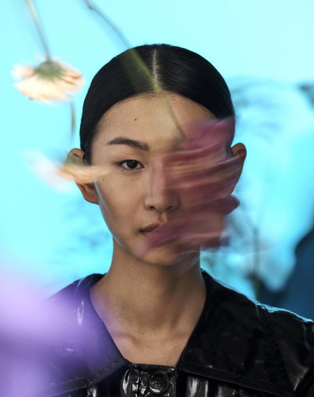 Portrait of an Asian woman with black hair and a black shiny jacket, with blurry flowers obscuring part of her face, against a blue background.