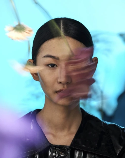 Portrait of an Asian woman with black hair and a black shiny jacket, with blurry flowers obscuring part of her face, against a blue background.