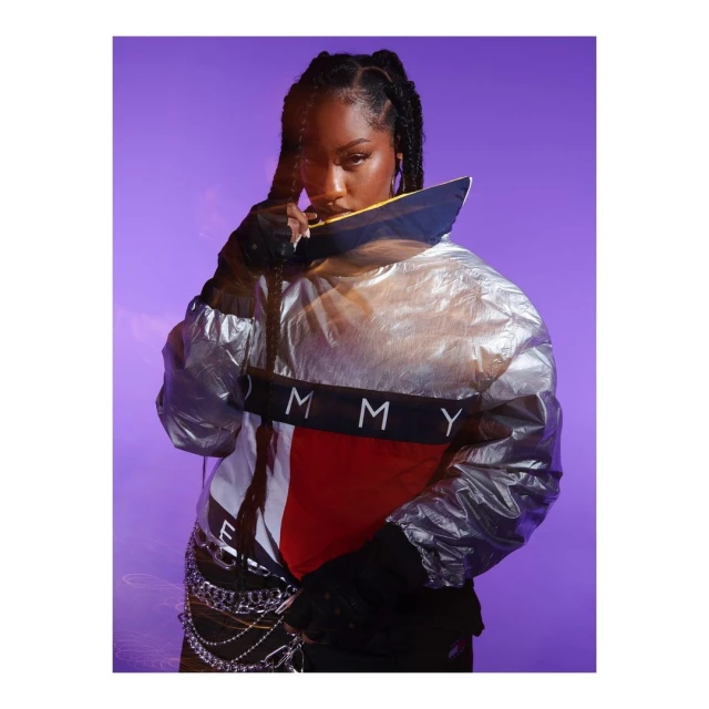 Portrait of a young woman with braids wearing a metallic silver jacket with red, white, and blue color blocking against a purple backdrop.