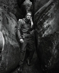 Man wearing a hooded jacket and backpack hiking through a narrow rocky canyon crevice in black and white