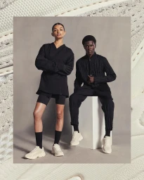 Two models standing and seated in black clothing and white sneakers against a grey backdrop, photographed by Mads Perch.