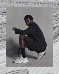 Black man crouching in black coat and shorts wearing grey sneakers against a textured grey studio background