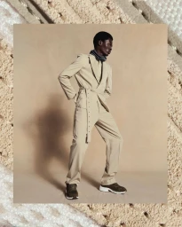 Black man standing in a beige belted suit and dark sneakers against a textured background by photographer Mads Perch.