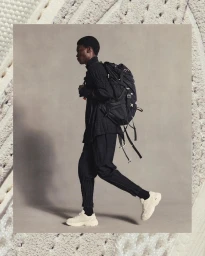 Black man wearing black clothing and a large black backpack walking against a grey backdrop