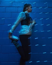 Female runner stretching leg against blue brick wall with glowing light trails in portrait composition