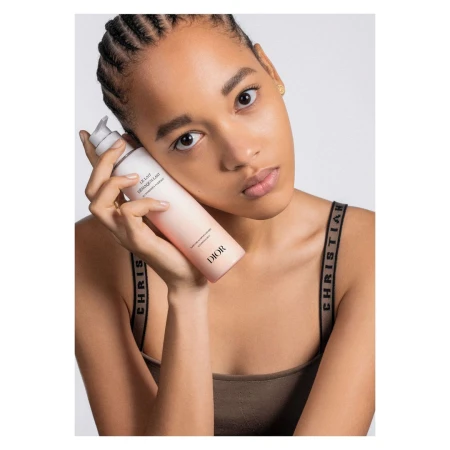 Tan-skinned woman poses in a close-up studio shot holding a Dior cleansing milk product against a bright neutral background.