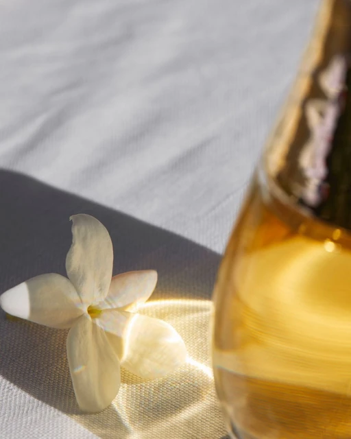 Close-up of a white jasmine flower next to a glass bottle with golden liquid, casting light on a wrinkled white fabric surface.