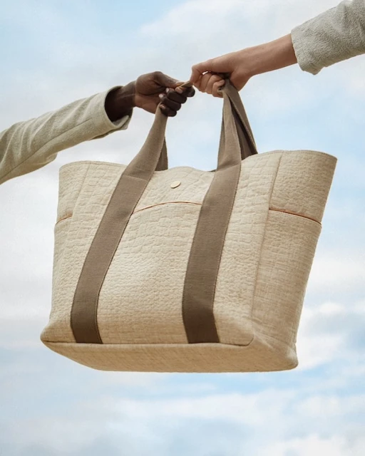 Two hands, one darker and one lighter, hold a textured beige tote bag with brown straps against a cloudy blue sky background.