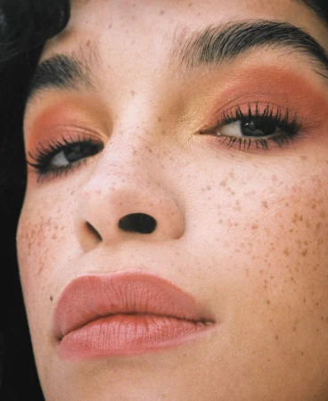 Close-up beauty portrait of a person with heavy freckles, warm terracotta eyeshadow, and natural brows by photographer Maeva Delacroix