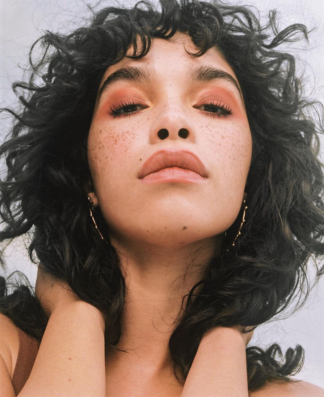 Close-up portrait of a young adult female with dark curly hair and bronze makeup holding her neck against a white background.