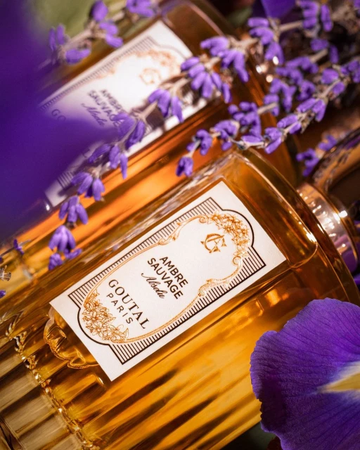 Close-up of two Goutal Paris Ambre Sauvage Absolu fragrance bottles with lavender and purple flower accents, shot in warm lighting.